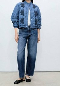 Niebieska pikowana kurtka bomberka z czarną floralną haftem, zestawiona z luźnymi niebieskimi dżinsami i czarnymi butami wsuwanymi.
