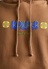 Smeđa dukserica s grafičkim logotipom koji prikazuje "KIDSUPER" u plavoj boji i "FOOTBALL CLUB" u zelenoj, ukrašena okruglim cvjetnim dizajnima u žutoj boji.