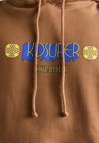 Hoodie marron avec un logo graphique portant "KIDSUPER" en bleu et "FOOTBALL CLUB" en vert, rehaussé de motifs floraux ronds en jaune.