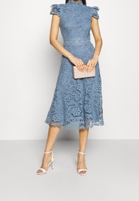 Niebieska koronkowa sukienka z wysokim kołnierzem i krótkimi falbaniastymi rękawami, z rozkloszowanym dołem. W zestawieniu z jasnoróżową kopertówką i białymi sandałkami na wysokim obcasie.