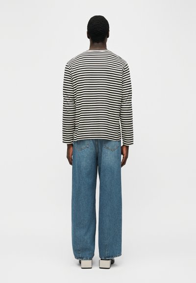 Άνδρας που στέκεται με την πλάτη προς τα εμπρός, φορώντας μακρυμάνικο πουκάμισο με ασπρόμαυρες ρίγες, φαρδιά μπλε τζιν και άσπρα παπούτσια.