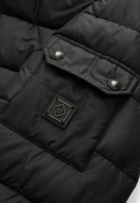 Sort pufferjakke-sektion med quiltet design, glat tekstur, trykknapper og et kvadratisk logo patch med et emblem.