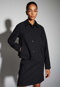 Zwarte op maat gemaakte jas met een kraag en knopen aan de voorkant, gecombineerd met een bijpassende rok. De stof lijkt glad met een gestructureerde pasvorm.