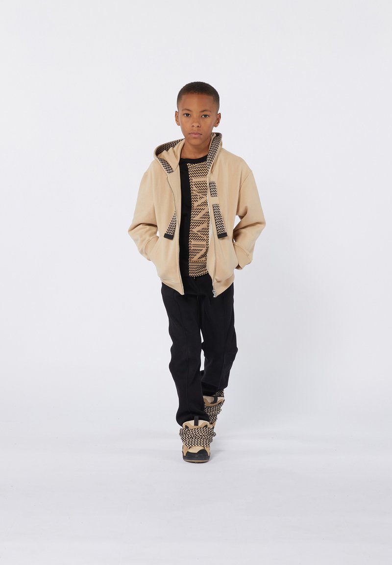 Haut à capuche beige zippé avec capuche à motifs et accents, chemise noire, pantalons noirs et chaussures présentant un design tissé avec des éléments de couleur beige.