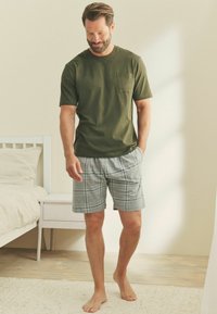 Olivenfarbene Kurzarm-T-Shirt mit einer Brusttasche, kombiniert mit grauen Karoshorts. Die Person steht barfuß auf einem hellen Boden.