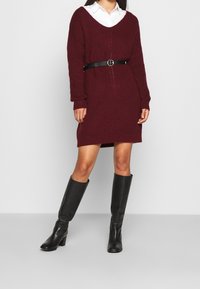 Robe tricotée bordeaux avec un décolleté en V, des manches longues et une ceinture noire à la taille. S'accorde avec des bottes en cuir noir jusqu'aux genoux.