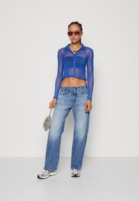 Cropped, long-sleeve, sheer blue button-up top, light blue distressed jeans, silver sneakers, and a metallic handbag.