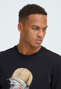 Sort t-shirt med korte ærmer med et grafik af en figur iført en beige kasket med et kranie og korslagte knogler. Blød tekstur, rund hals.