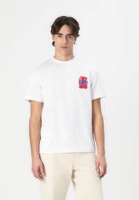 White cotton t-shirt with short sleeves featuring a red and blue graphic design on the left chest area. Plain back, relaxed fit.