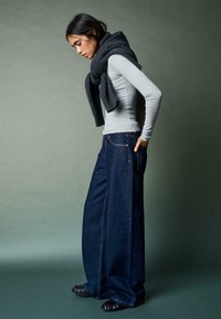 Ženska v širokih temno modrih kavbojkah, svetlo sivi majici z dolgimi rokavi in temnem puloverju, premeščenem čez ramena, stoji bočno z rokami v žepih.