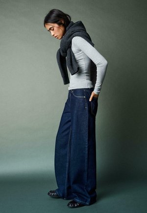Žena nosi široké tmavomodré džínsy, svetlosivé tričko s dlhým rukávom, tmavý sveter prehodený cez ramená, stojí bokom s rukami v záhyboch nohavíc.