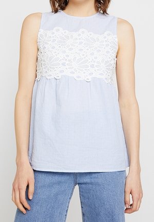 Bezrękawnik w jasnoniebieskim kolorze z białym koronkowym panelem w kwiaty na wysokości klatki piersiowej, noszony z niebieskimi dżinsami średniego odcienia na osobie.
