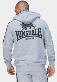 Šedá mikina s kapucňou s grafikou leva a textom "LONSDALE LONDON" na chrbte. Obsahuje kapucňu so šnúrkou a rebrované manžety.