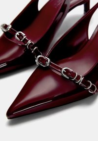 Talons escarpins en cuir verni bordeaux avec des bouts pointus et une finition brillante. Présente un accent en chaîne argentée sur la bride.