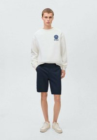 Vit sweatshirt med en blå våg-logotyp på bröstet, kombinerad med mörkblå shorts och beige sneakers. En enkel, avslappnad design.