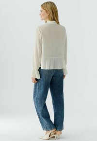 Woman wearing sheer cream blouse with ruffled cuffs and hem, blue straight-leg jeans, and cream pointed-toe kitten heels, standing facing sideways.