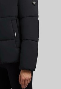 Manica e tasca laterale di una giacca a vento nera con patch logo "Khujo" sull'avambraccio, indossata da una persona con la mano visibile e pantaloni neri.