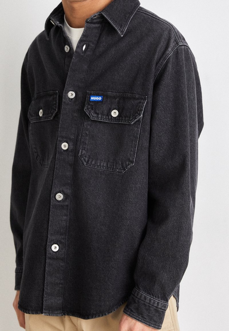 Homme portant une chemise noire à boutons avec deux poches poitrine, dont une étiquetée "HUGO,", par-dessus un sous-vêtement blanc et un pantalon beige.