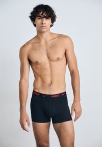 Schwarze Boxershorts mit einem roten Nike-Logo am Bund; Modell ist oben ohne und zeigt eine definierte Physik vor neutralem Hintergrund.