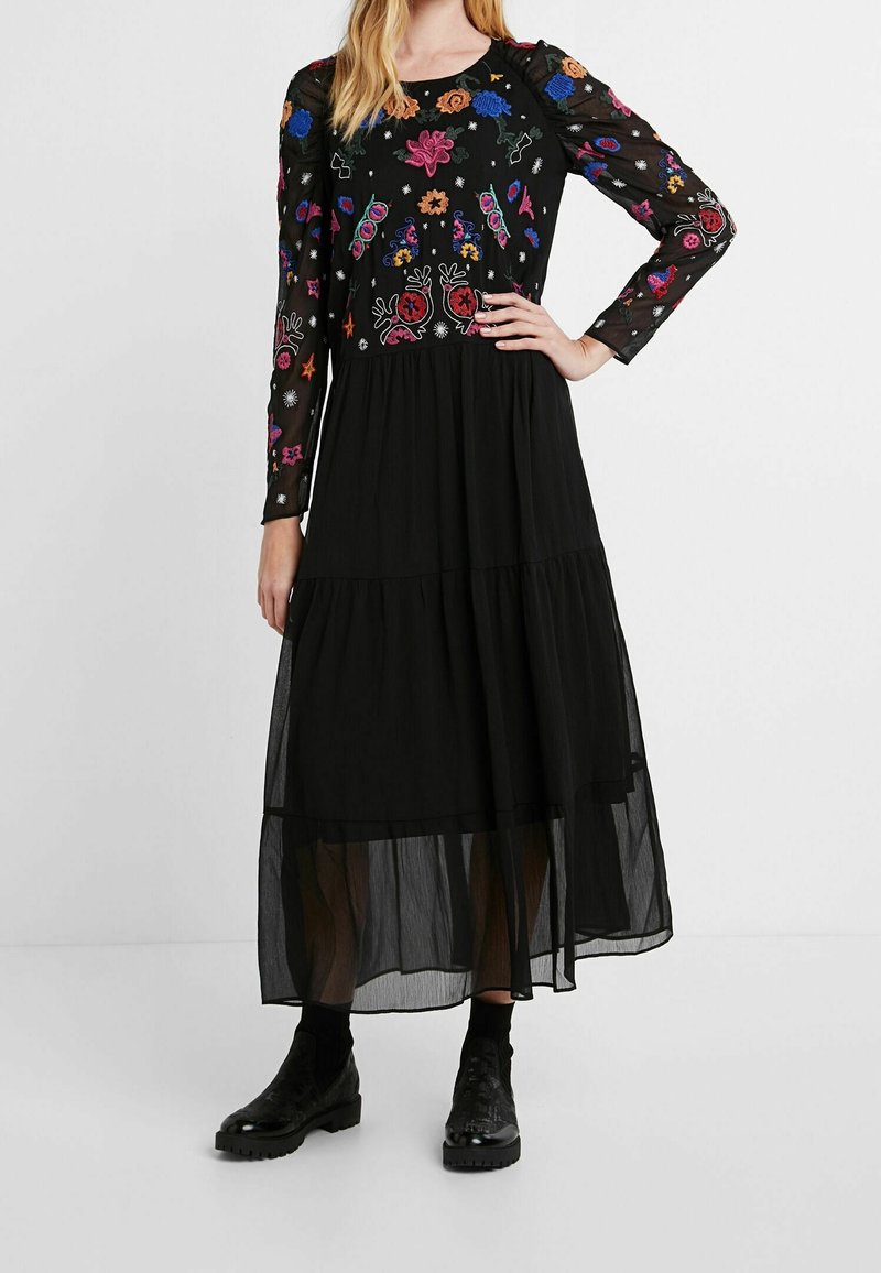 Schwarzes Maxikleid mit langen, transparenten Ärmeln, das farbenfrohe Blumenstickereien auf dem Oberteil aufweist, ein geschichtetes Design hat und einen fließenden, transparenten Rock besitzt. Schwarze Stiefel.