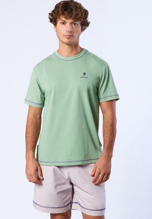 Giovane uomo con t-shirt a maniche corte verde chiaro e pantaloncini beige chiaro, in piedi davanti a uno sfondo chiaro e semplice.