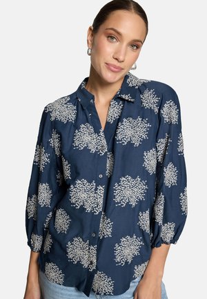 Femme portant un chemisier bleu marine à boutons avec des motifs floraux blancs et des manches trois-quarts, associé à un jean bleu clair.