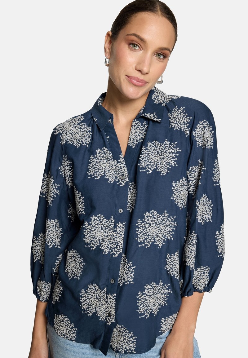 Femme portant un chemisier bleu marine à boutons avec des motifs floraux blancs et des manches trois-quarts, associé à un jean bleu clair.