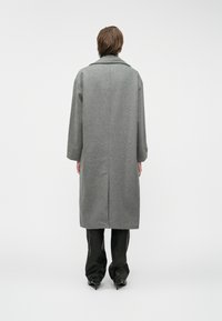 Asmuo stovi nukreipęs nugarą į žiūrovą, apsirengęs ilgu pilku paltu, juodomis kelnėmis ir juodais aukštakulniais bateliais priešais paprastą foną.