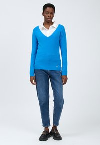 Blauwe v-hals trui van gebreid materiaal, gecombineerd met een wit overhemd met kraag en blauwe jeans. De look wordt compleet gemaakt met zwarte instappers.