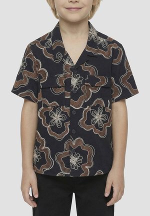 Niño con una camisa negra de manga corta con grandes estampados florales marrones y blancos y bolsillos en el pecho delanteros.