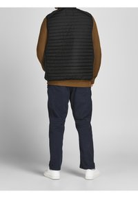 Czarna pikowana kamizelka na brązowym swetrze o fakturowanej strukturze, zestawiona z ciemnoniebieskimi spodniami i białymi trampkami. Kamizelka ma stojący kołnierz i zamek błyskawiczny.