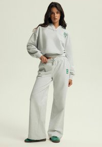 Jasnoszara bluza z dekoltem w serek i zielonym detalem logo, w zestawie z pasującymi spodniami dresowymi o szerokich nogawkach z elastycznym pasem i zielonym akcentem.