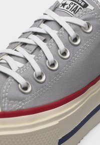 Κοντινή λήψη γκρι πάνινο αθλητικό παπούτσι με λευκά κορδόνια, ασημένια μεταλλικά ρούθουνια, κόκκινη ρίγα στη μέση της σόλας και υφασμένη μπεζ εξωτερική σόλα.