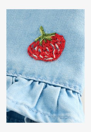 Šviesiai mėlynas džinsas su raukšlėtu kraštu, puošiamas detaliai išsiuvinėtomis raudonomis braškėmis su žaliomis detalėmis, ant lygaus tekstūrinio fono.