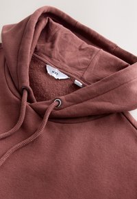 Sweat à capuche marron avec une texture douce, des cordons de serrage, et une poche kangourou. Caractérisé par un intérieur confortable et une étiquette portant la mention "NXT".