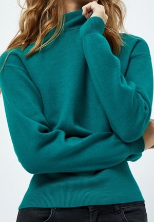 Femme portant un pull à manches longues couleur sarcelle avec des poignets et un ourlet côtelés, posant avec une main près du menton sur un fond clair uni.