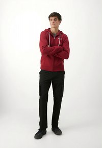 Casaco com fecho de correr vermelho com cordões brancos, combinado com calças cargo pretas com dois bolsos laterais e sapatos pretos. Design simples e funcional.