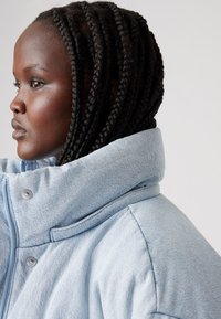 Light blue denim oversized jacket with a high collar, metallic snaps, and structured shoulder seams. Features prominent stitching detail.