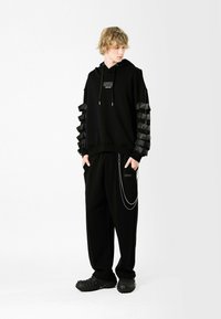 Sudadera negra con mangas superpuestas y acanaladas; presenta un bolsillo frontal y capucha con cordón. Combinada con pantalones negros holgados y zapatos texturizados.