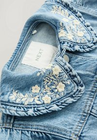 Col de vêtement en denim bleu clair avec broderie florale blanche et bordure tressée, montrant une étiquette de marque "NEXT" blanche à l'intérieur.