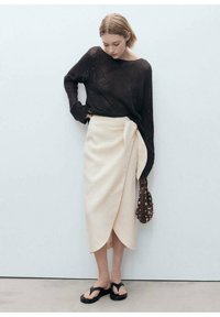 Zwarte gebreide trui, crèmekleurige wikkelrok, bruine etui-tas en zwarte slippers. Minimalistisch ontwerp, zachte texturen en neutrale kleuren.