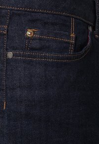 Jeans de mezclilla azul oscuro con costuras cobre, con cierre de botón y un pequeño logotipo en el bolsillo delantero. Textura suave, corte clásico.