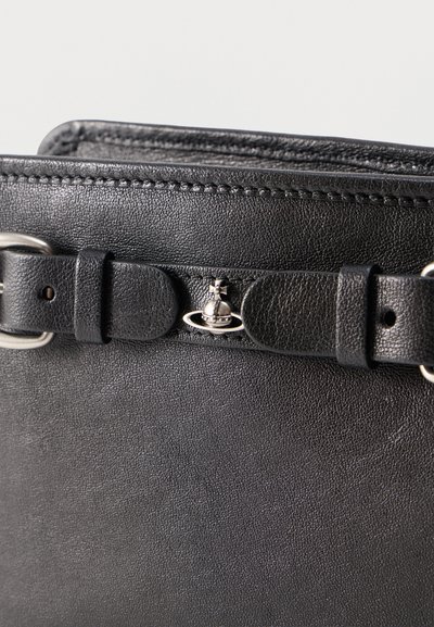 Κοντινή λήψη μαύρης δερμάτινης τσάντας που δείχνει υφή, ραμμένα άκρα, λεπτομέρεια ζώνης και ένα μικρό ασημί μεταλλικό λογότυπο σε σχήμα Κρόνου.