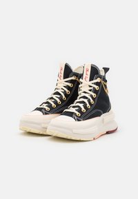Sneakers alte con sopra in tela nera, lacci e suola color crema, occhielli dorati e un accento decorativo di catena dorata.