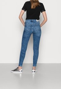 Jeans ajustados de mezclilla azul con una textura desgastada, de cintura alta y con dos bolsillos traseros. Combinados con una camiseta negra y zapatillas plateadas.