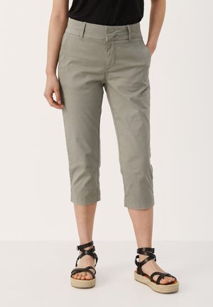 Kvinde iført lysegrå, korte bukser og sorte sandaler med stropper og plateauhæl, stående med den ene hånd i lommen.