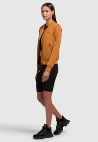 Oranje bom jacket met ribgebreide manchetten en kraag, voorzien van een ritssluiting aan de voorkant en zijzakken, gecombineerd met zwarte shorts en sportschoenen.