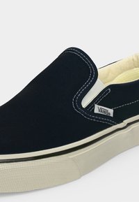 Μαύρο παπούτσι Vans slip-on με λευκό ράψιμο, λευκές ελαστικές πλευρικές λεπτομέρειες και λευκή λαστιχένια σόλα με υφή λαβής στο δάχτυλο.