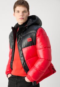 Κόκκινη και μαύρη φούτερ ζακέτα με κουκούλα, με ραφές, γυαλιστερή υφή και μικρή ετικέτα λογότυπου στο στήθος. Ελαστικοί καρποί και στρίφωμα.