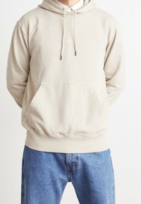 Beige huvtröja med framficka, ribbade mudd och fåll, samt snören med silverändar. Matchas med blå denimjeans.